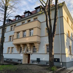 NOW BUD NOWAKOWSKI PAWEŁ - Odnowiona elewacja kamienicy z balkonem w odcieniach szarości i beżu, widoczna z poziomu ulicy, z drzewami po bokach, w słoneczny dzień.