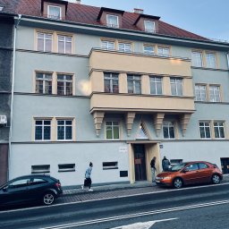 NOW BUD NOWAKOWSKI PAWEŁ - Kamienica po renowacji z balkonem w odcieniach beżu i szarości. Dwie osoby wchodzą do budynku. Na pierwszym planie zaparkowane samochody.