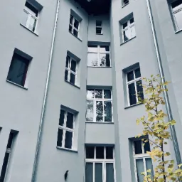 Jasnoszara elewacja budynku wielorodzinnego z licznymi oknami, widziana z dołu pod kątem, z żółknącym drzewkiem na pierwszym planie po prawej stronie.