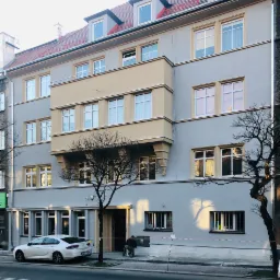 Szara kamienica z beżowym balkonem i czerwoną dachówką, w trakcie prac malarskich przy wejściu, zabezpieczona taśmą ostrzegawczą, z zaparkowanym białym samochodem i drzewami bez liści.