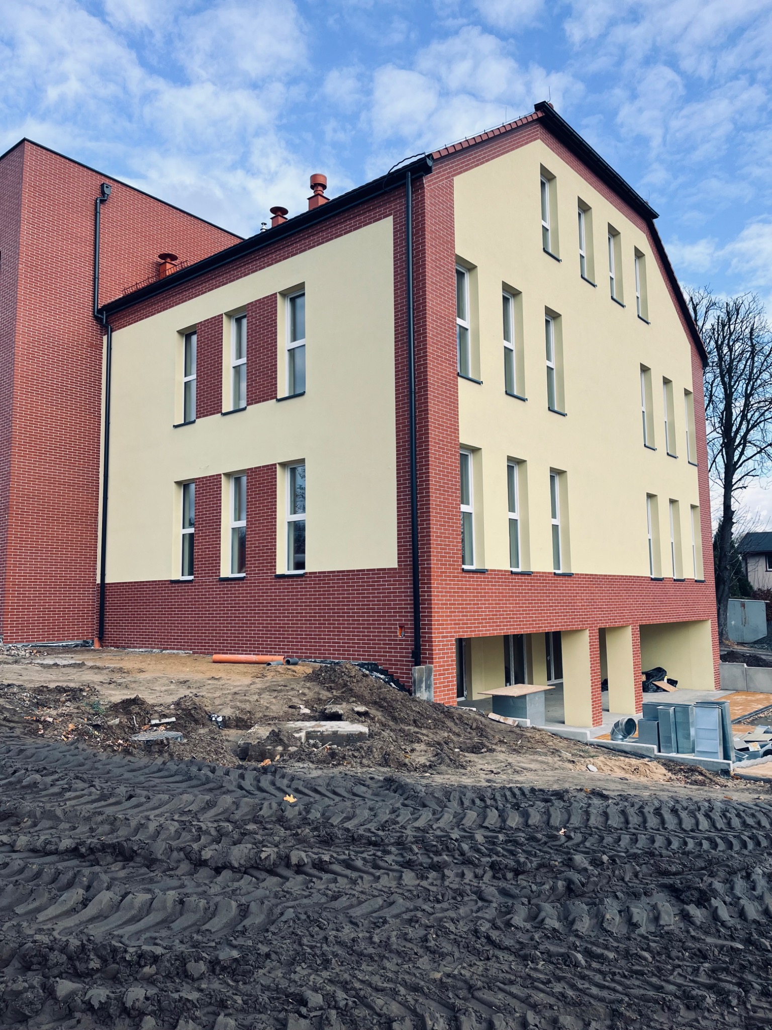 Nowoczesny budynek z elewacją w odcieniach beżu i cegły. Widoczne białe ramy okienne, ciemna linia dachu i przygotowany teren wokół budynku. Słoneczny dzień.