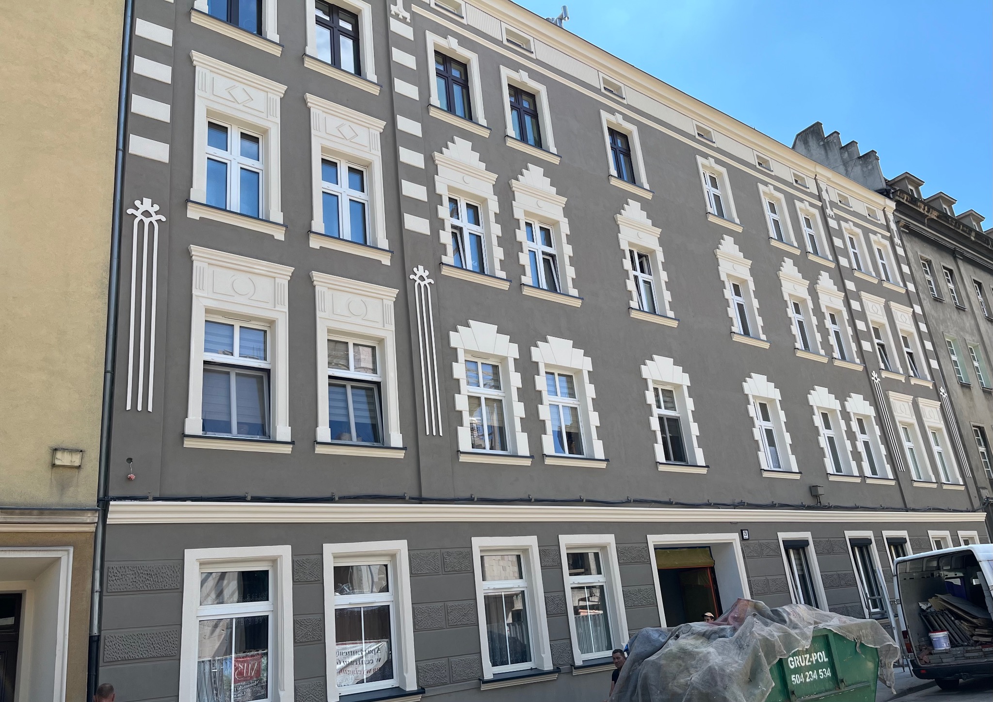 Szara kamienica po renowacji elewacji z dekoracyjnymi białymi obramowaniami okien. Kontener na gruz i częściowo widoczny samochód dostawczy.