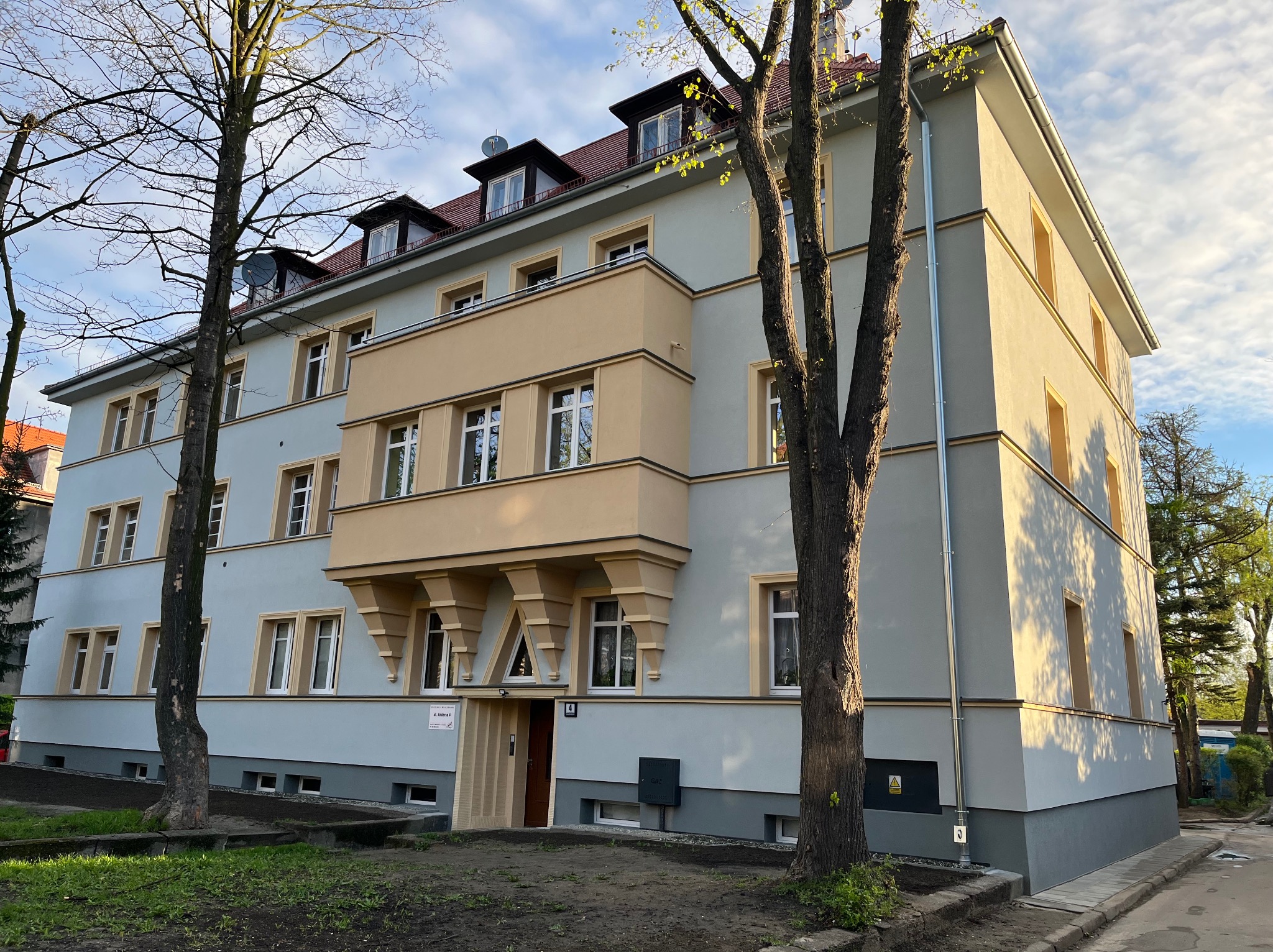 Odnowiona elewacja kamienicy z balkonem w odcieniach szarości i beżu, widoczna z poziomu ulicy, z drzewami po bokach, w słoneczny dzień.