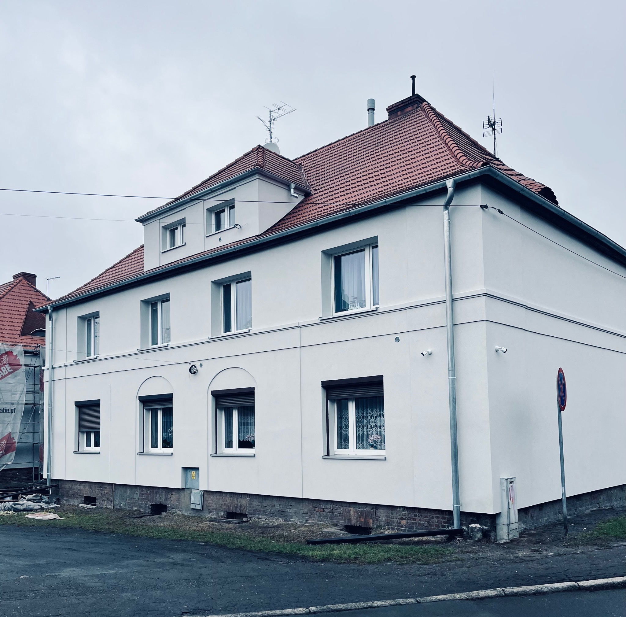 Dwupiętrowy dom z czerwoną dachówką po renowacji elewacji. Widoczne detale architektoniczne, czyste białe ściany i odnowiona fasada. W tle rusztowania przy sąsiednim budynku.