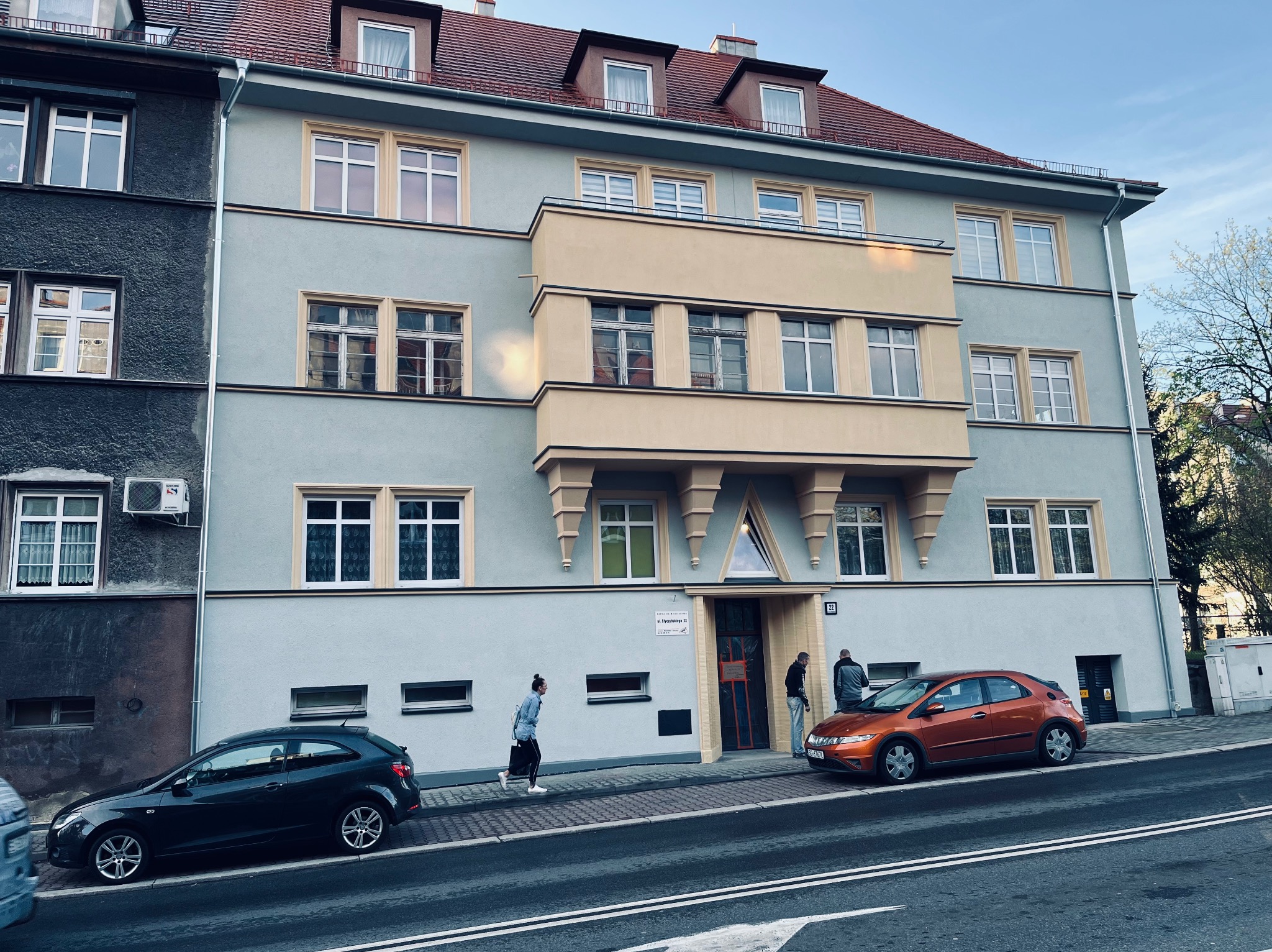 Kamienica po renowacji z balkonem w odcieniach beżu i szarości. Dwie osoby wchodzą do budynku. Na pierwszym planie zaparkowane samochody.