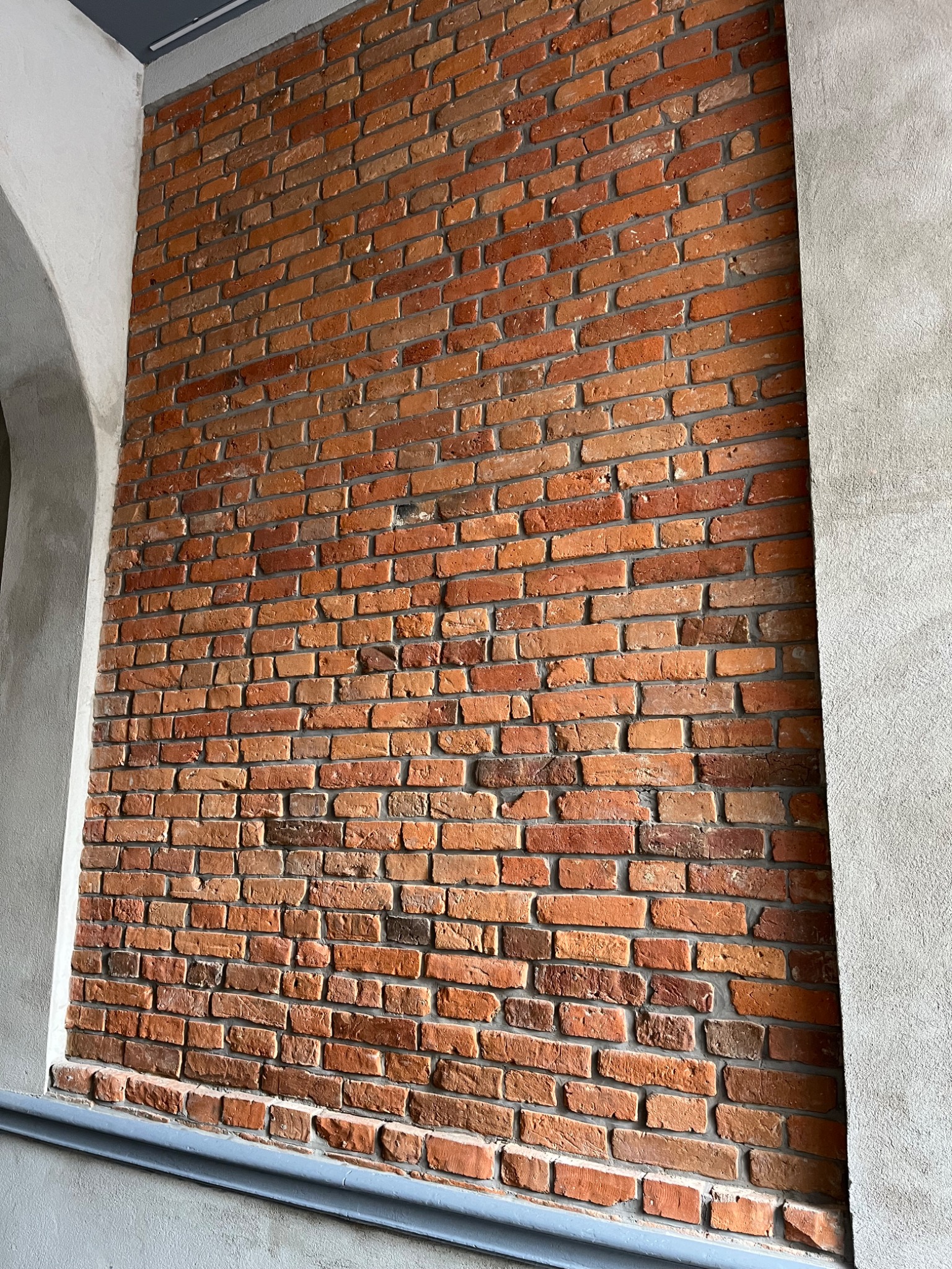 Ceglana ściana wewnętrzna z szarą ramą okienną i listwą przypodłogową. Widoczna faktura cegieł i spoin, surowy, industrialny styl wykończenia wnętrza.