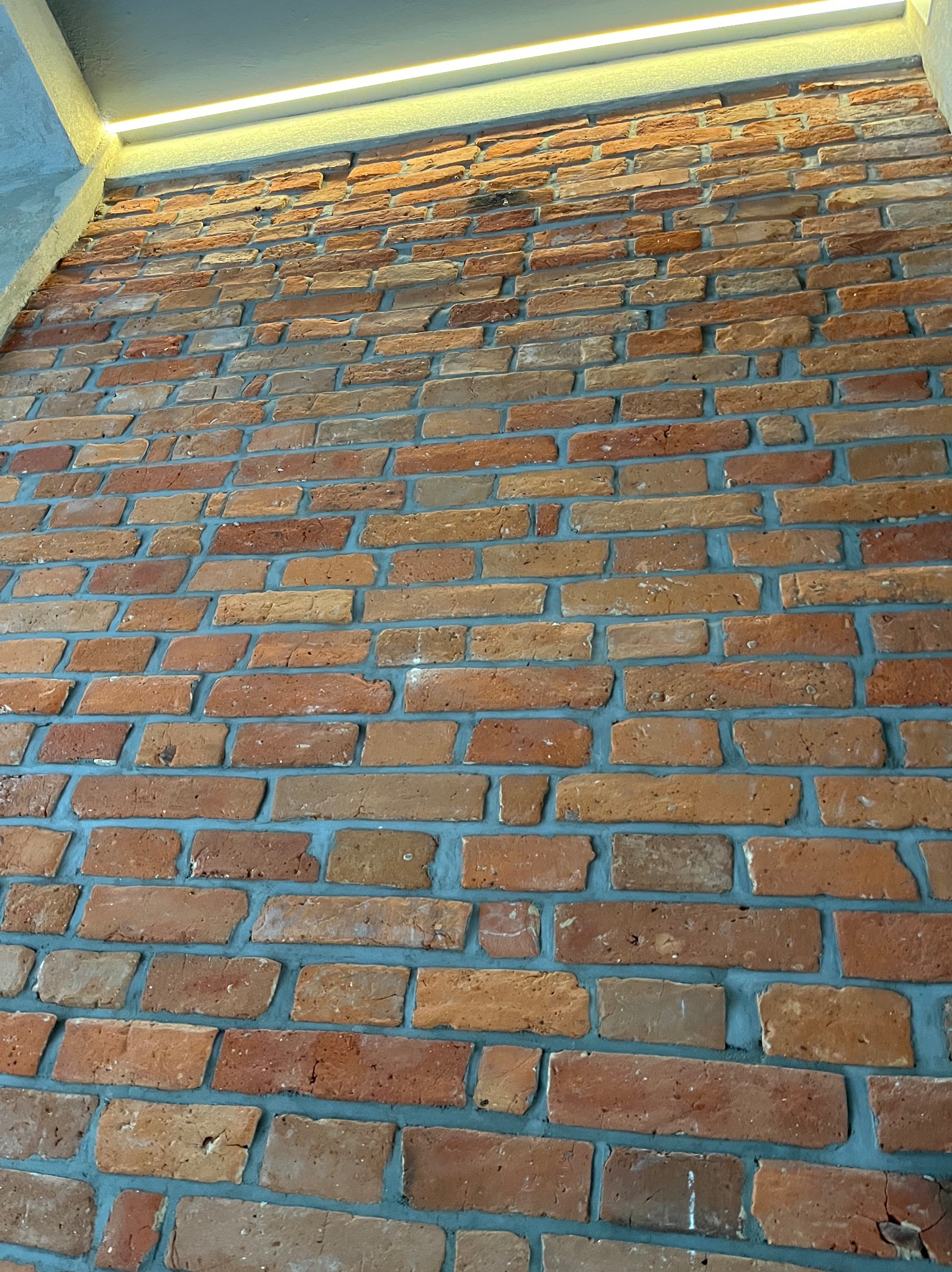 Ceglana ściana wewnętrzna z szarą fugą i oświetleniem LED na górze. Widok z dołu podkreśla fakturę i kolorystykę cegieł.