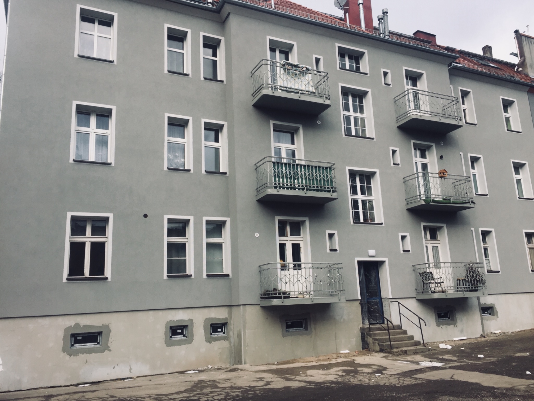 Odnowiona elewacja kamienicy w szarym kolorze z białymi ramami okiennymi i balkonami z metalowymi balustradami, widoczne ślady prac wykończeniowych przy oknach piwnicznych.