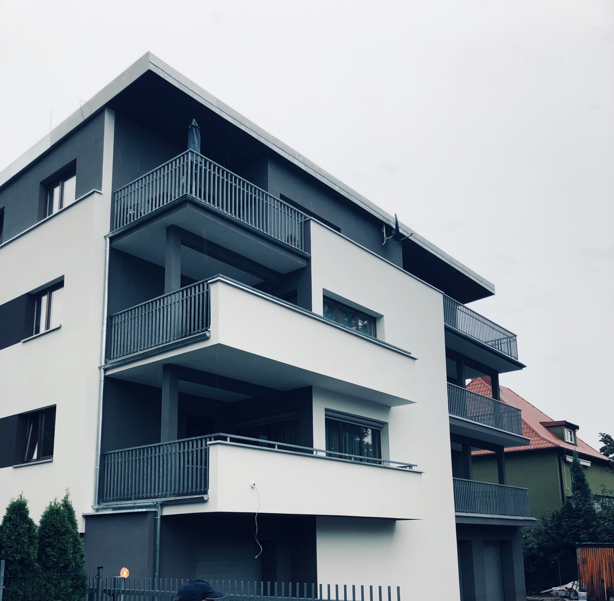 Nowoczesny budynek wielorodzinny z balkonami wykończony w bieli i szarości, widok z dołu na elewację i fragment otoczenia.