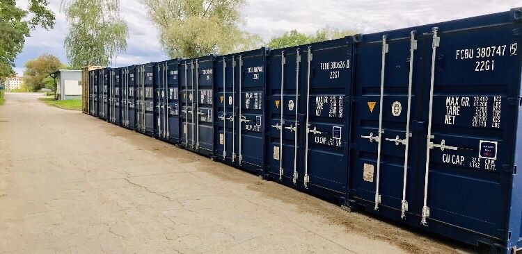 Szereg ciemnoniebieskich kontenerów ustawionych jeden za drugim na betonowym placu, widoczne oznaczenia i numery identyfikacyjne na kontenerach, w tle drzewa i budynek.