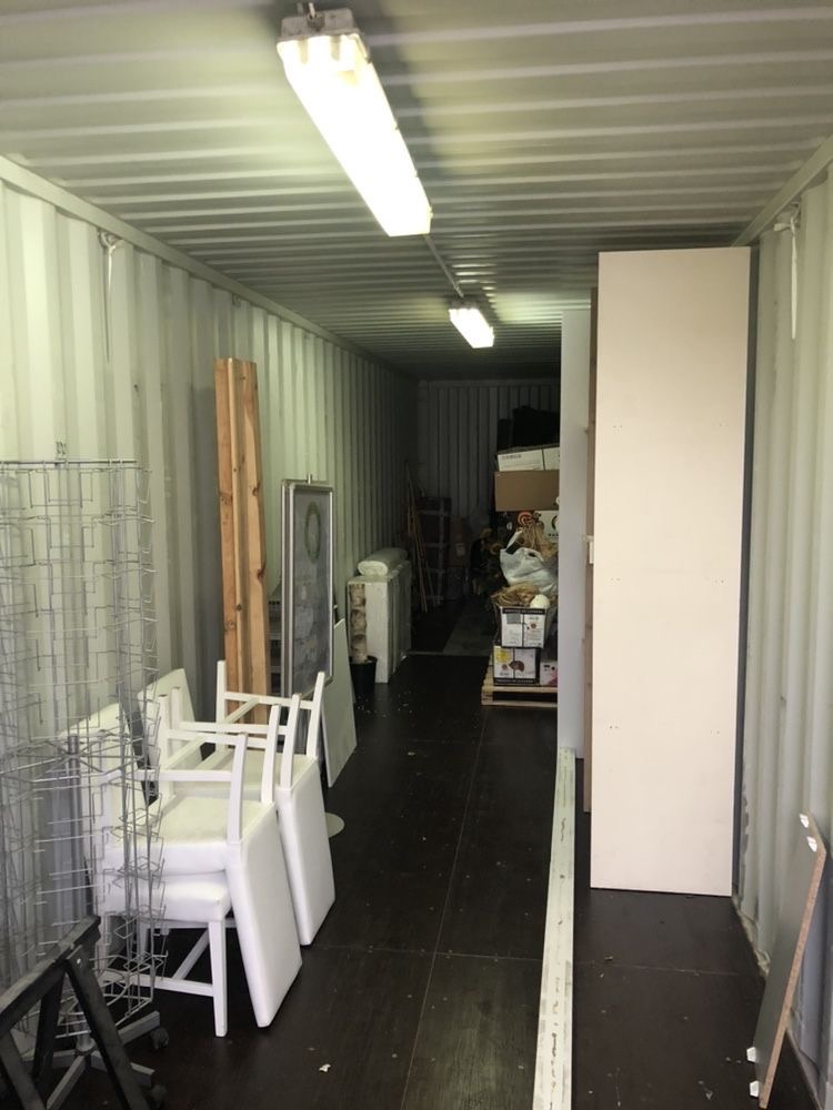 Wnętrze kontenera magazynowego oświetlonego lampami jarzeniowymi, wypełnione różnorodnymi przedmiotami, w tym stertą białych krzeseł, metalowymi regałami i kartonami w głębi.
