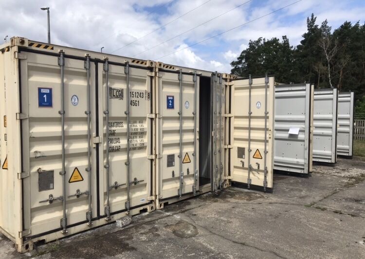 Rząd beżowych kontenerów magazynowych, jeden z otwartymi drzwiami, widoczne oznaczenia i numery identyfikacyjne, tło z drzewami i błękitnym niebem.