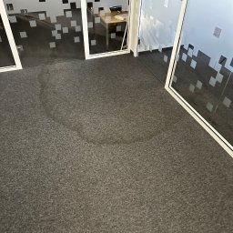 XCLEANERS - Szary dywan biurowy z widocznym śladem po plamie, częściowo wyczyszczony, w tle szklane ściany działowe z motywem pikseli i logo firmy Omega Group.