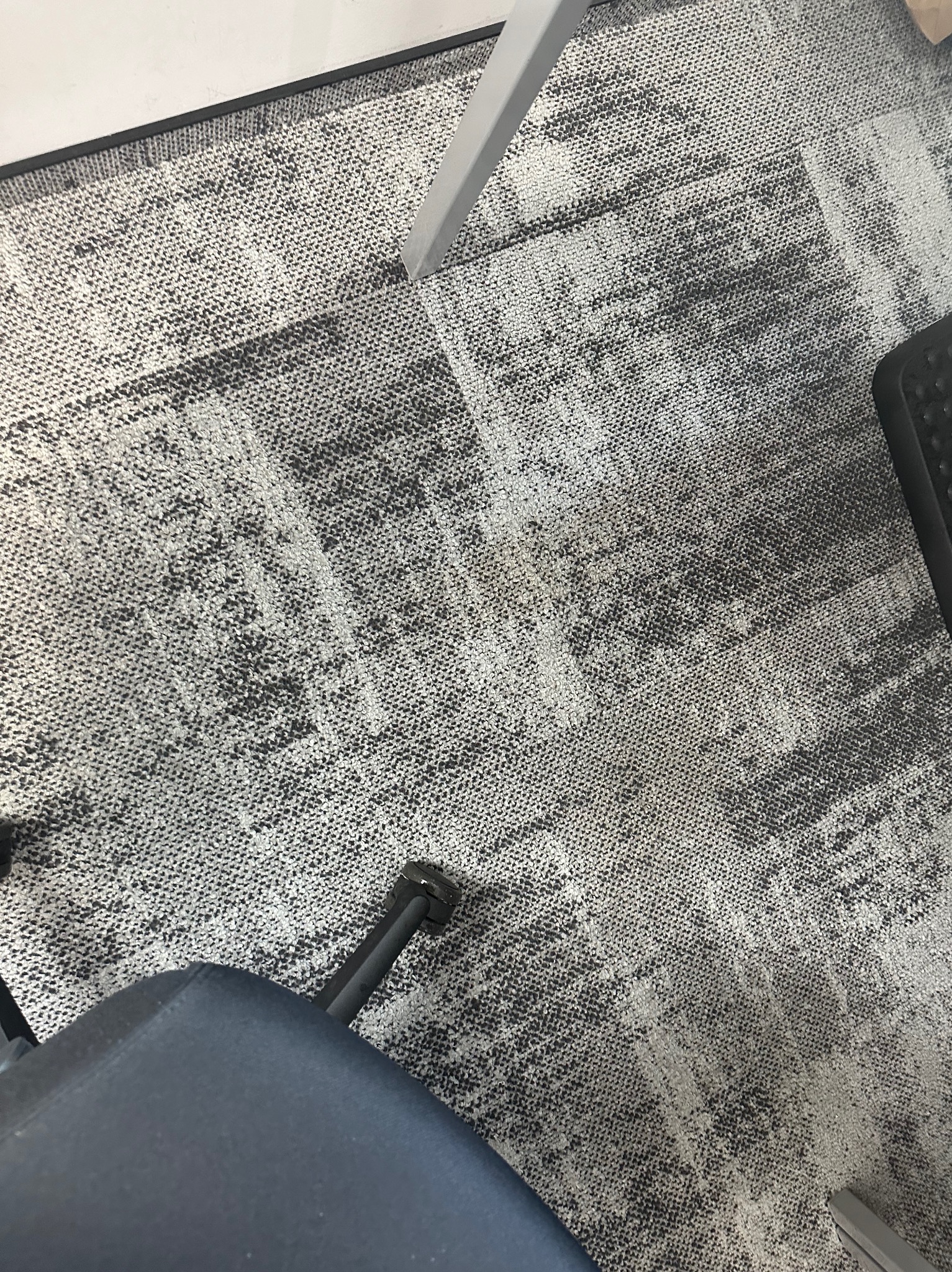 Wzór dywanu biurowego w odcieniach szarości z widocznymi śladami użytkowania i fragmentami mebli biurowych, w tym nogi krzesła i fragmentu biurka.