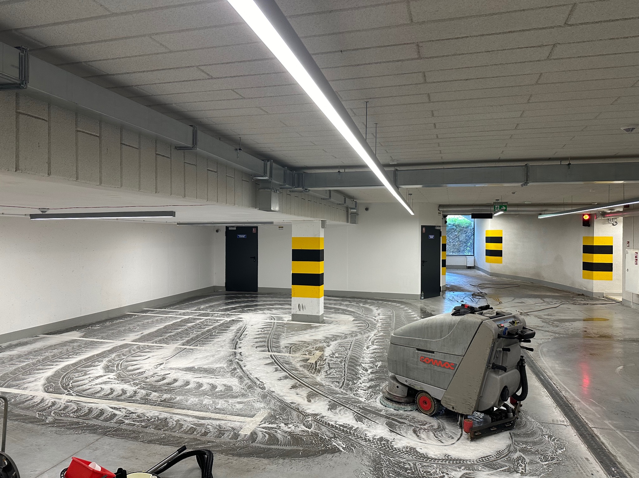 Szorowarka automatyczna w trakcie czyszczenia posadzki w garażu podziemnym, widoczne ślady detergentu, słupy oznaczone pasami ostrzegawczymi.