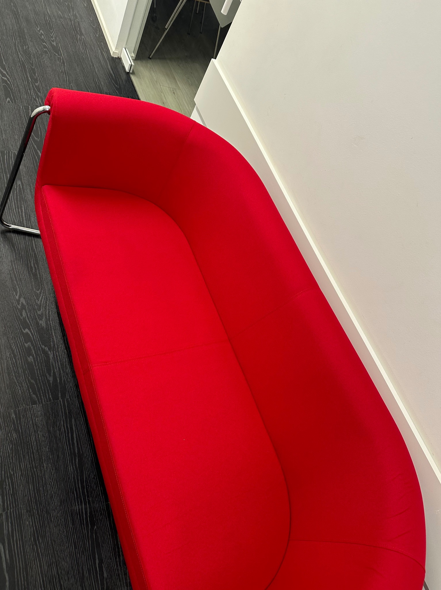 Czerwona, nowoczesna kanapa z metalowymi nogami, ustawiona pod kątem w biurze na tle ciemnej podłogi i białej ściany, widok z góry.