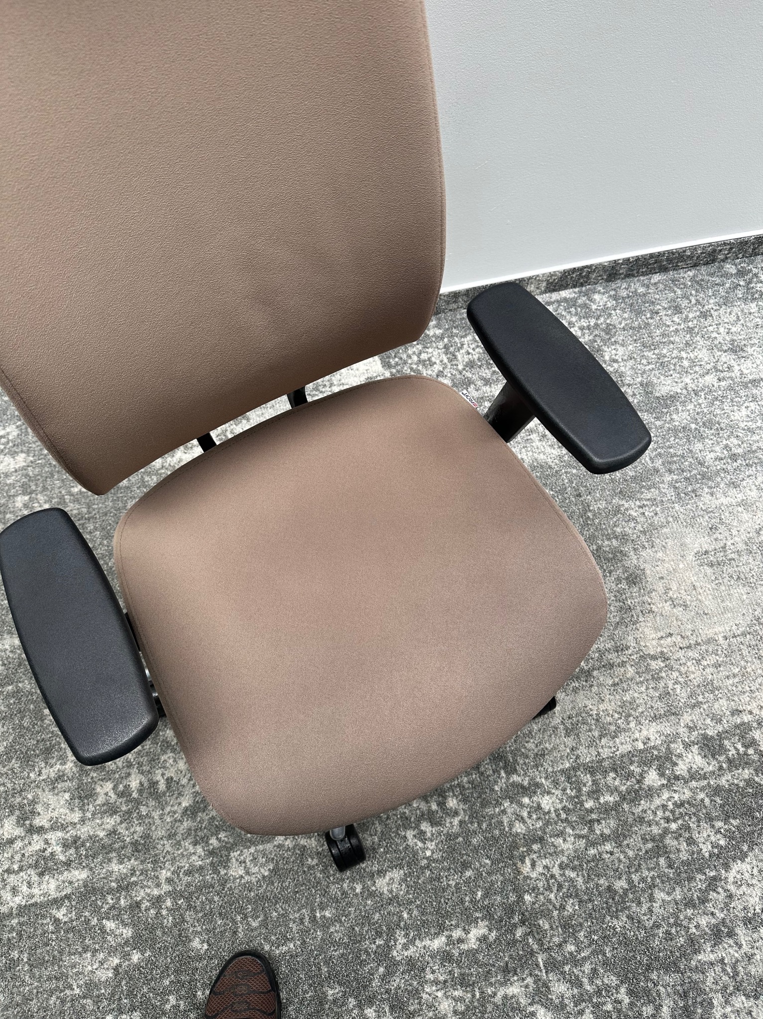 Beżowe krzesło biurowe z czarnymi podłokietnikami, widok z góry na siedzisko i oparcie, na tle szarej wykładziny dywanowej. Widoczny fragment buta w rogu.
