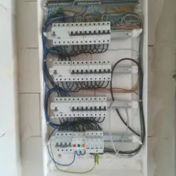 Otwarta skrzynka rozdzielcza z zainstalowanymi bezpiecznikami i przewodami w surowym wnętrzu, widoczne ślady po montażu na ścianie z betonu komórkowego.