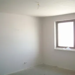 Wnętrze pokoju po nałożeniu tynku, widoczne gładkie, jasne ściany i sufit, okno z ciemną ramą i szary betonowy podkład pod posadzkę.