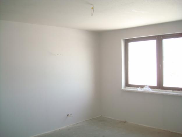 Wnętrze pokoju po nałożeniu tynku, widoczne gładkie, jasne ściany i sufit, okno z ciemną ramą i szary betonowy podkład pod posadzkę.
