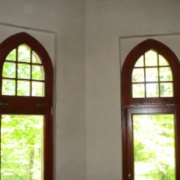 Dwa identyczne, drewniane okna w stylu gotyckim, z widokiem na zieleń, osadzone w ścianie pokrytej jasnym, gładkim tynkiem.