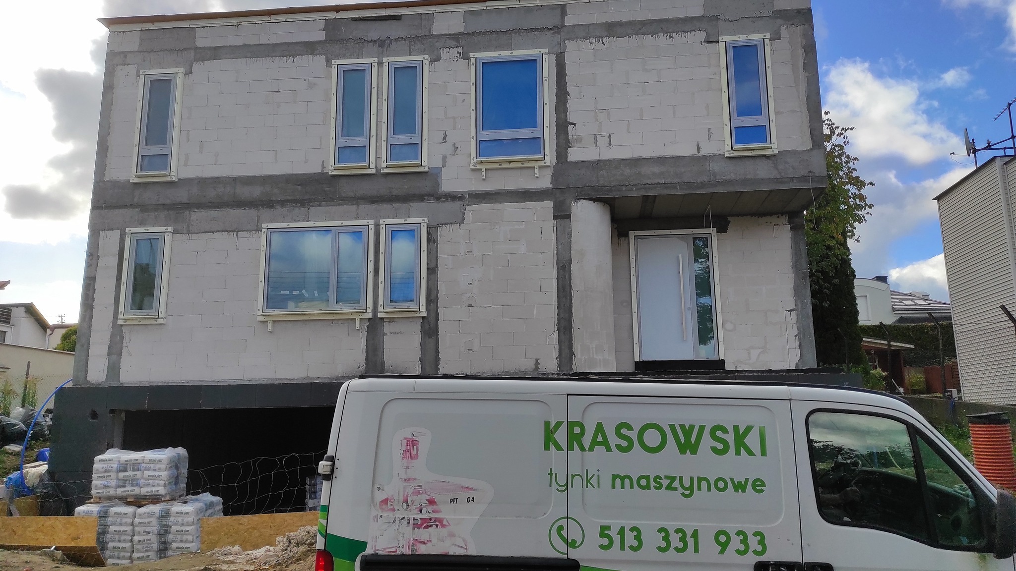 Surowy stan dwupiętrowego domu z białego bloczka, z częściowo osadzonymi oknami i widocznym zbrojeniem, z zaparkowanym przed nim busem firmy budowlanej z reklamą usług tynkarskich.