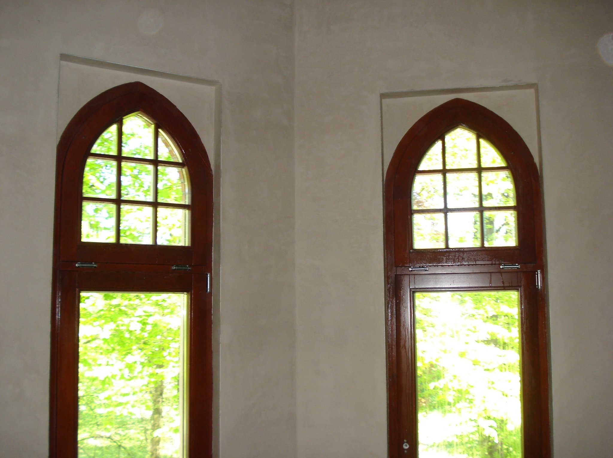 Dwa identyczne, drewniane okna w stylu gotyckim, z widokiem na zieleń, osadzone w ścianie pokrytej jasnym, gładkim tynkiem.