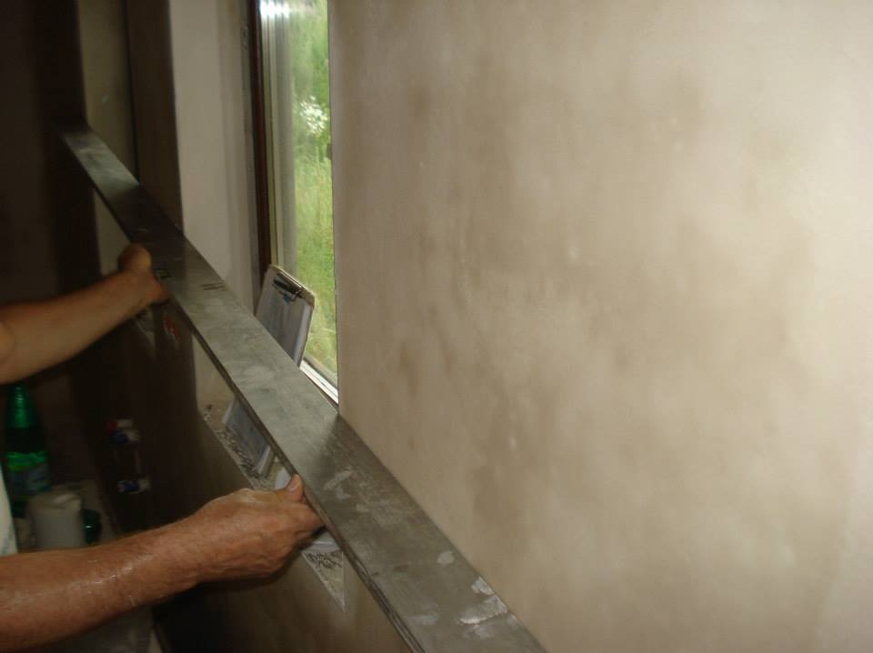Wyrównywanie świeżo nałożonego tynku na ścianie przy oknie za pomocą długiej, metalowej łaty.
