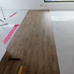 Fragment podłogi w trakcie układania paneli, widoczne narzędzia: młotek i poziomica, oraz folia izolacyjna na nieułożonej części podłogi.