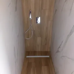 Kabina prysznicowa walk-in z odpływem liniowym, wyłożona płytkami imitującymi drewno i marmur, z chromowaną słuchawką prysznicową i baterią.