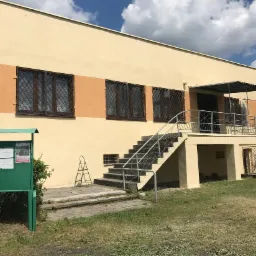 Dwukondygnacyjny budynek z beżową elewacją, pomarańczowym pasem i kratami w oknach, widoczne schody wejściowe i zieleń, częściowo zakryte folią zabezpieczającą.