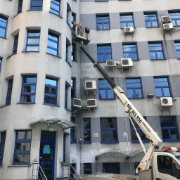 Pracownik w koszu podnośnika na tle szarej elewacji budynku z licznymi oknami i klimatyzatorami, widoczny numer telefonu na ramieniu podnośnika.