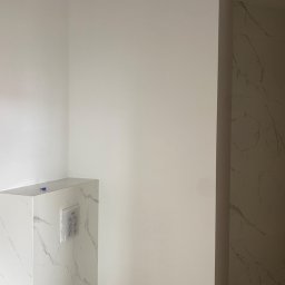 mykola katruk - Wykończona toaleta z zabudowaną spłuczką, wykończona płytkami imitującymi biały marmur, z widocznym przyciskiem spłukującym i fragmentem podłogi oraz ścianą pomalowaną na biało.