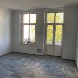 Wnętrze pokoju po malowaniu ścian na biało, z szarą wykładziną dywanową w geometryczne wzory, widoczne dwa okna i niezamaskowane gniazdka elektryczne.