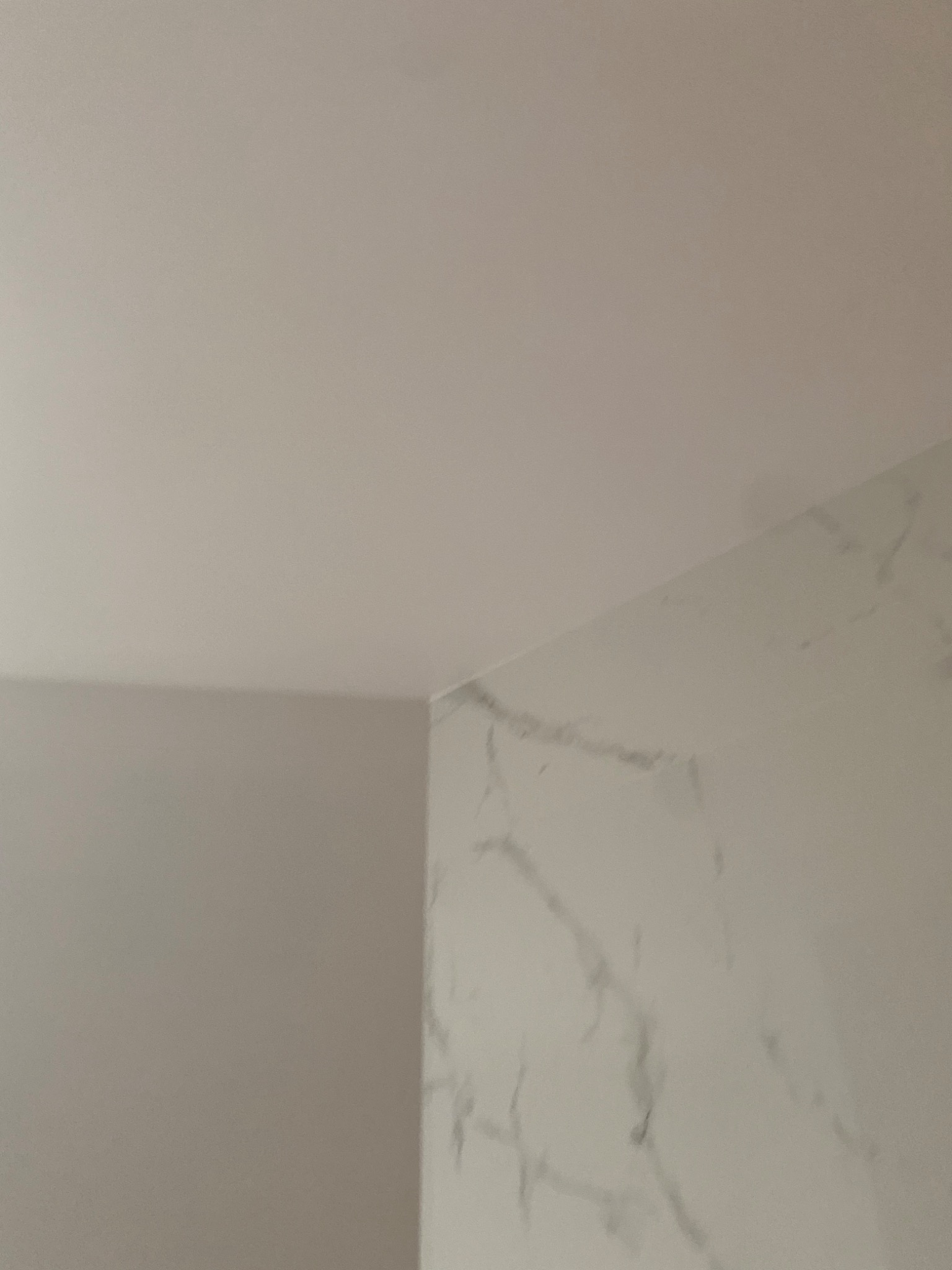 Gładki, pomalowany sufit w rogu pomieszczenia, sąsiadujący ze ścianą wykończoną płytami imitującymi biały marmur z szarymi żyłami.