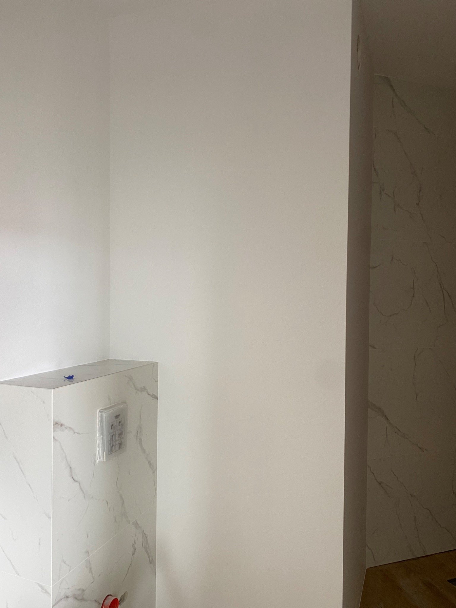 Wykończona toaleta z zabudowaną spłuczką, wykończona płytkami imitującymi biały marmur, z widocznym przyciskiem spłukującym i fragmentem podłogi oraz ścianą pomalowaną na biało.