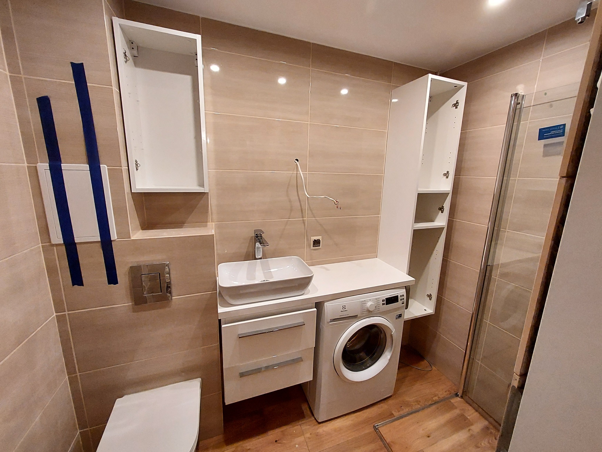 Nowoczesna łazienka w trakcie wykończenia, z beżowymi płytkami, białą umywalką nablatową, pralką Electrolux zabudowaną pod blatem i wiszącymi szafkami bez frontów, widoczne niepodłączone kable.