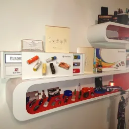 Półki z ekspozycją gadżetów reklamowych: pendrive'y z logo firm, wizytówki, notatniki i inne materiały promocyjne, umieszczone w nowoczesnych, białych półkach z czerwonym wnętrzem.