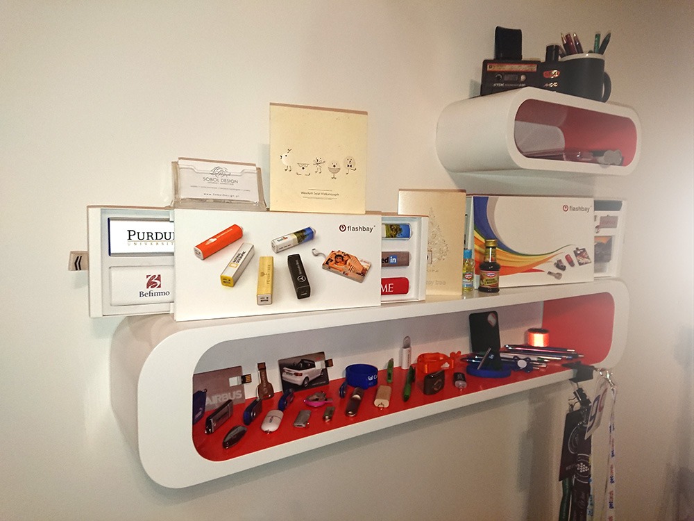 Półki z ekspozycją gadżetów reklamowych: pendrive'y z logo firm, wizytówki, notatniki i inne materiały promocyjne, umieszczone w nowoczesnych, białych półkach z czerwonym wnętrzem.