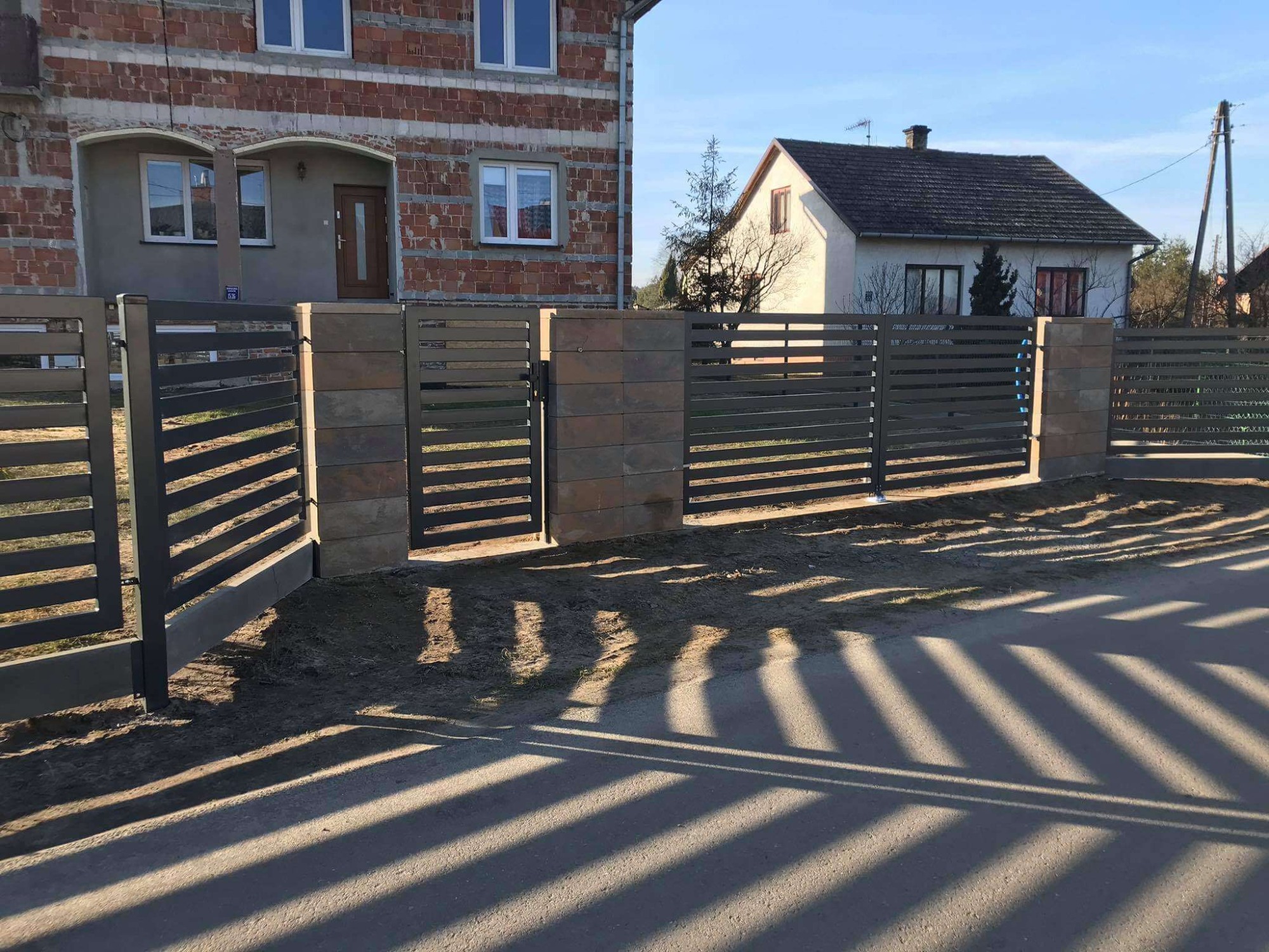 Nowoczesne ogrodzenie z poziomych, grafitowych profili metalowych i słupków z szarej kostki, na tle domu w trakcie budowy i drugiego, gotowego domu.