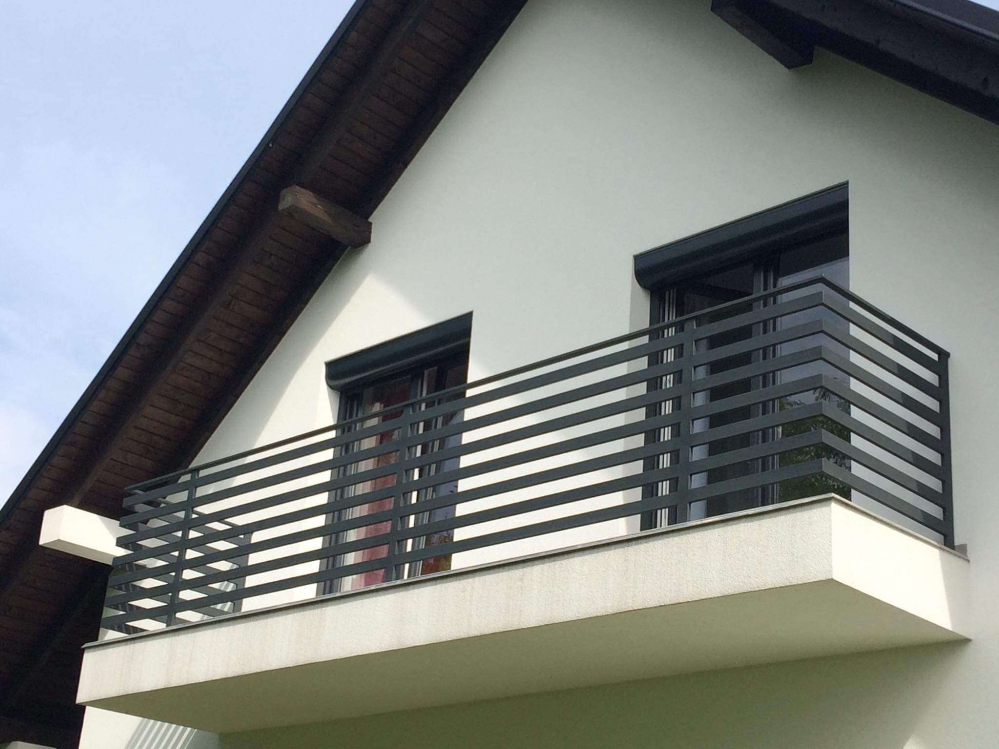Nowoczesna, pozioma, metalowa balustrada balkonowa w kolorze grafitowym, zamontowana na białym budynku z ciemnobrązowym dachem.