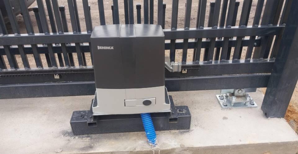 Zainstalowany napęd elektryczny Beninca do bramy przesuwnej, widoczna metalowa brama z pionowymi prętami, niebieski peszel kablowy, szara betonowa podstawa.