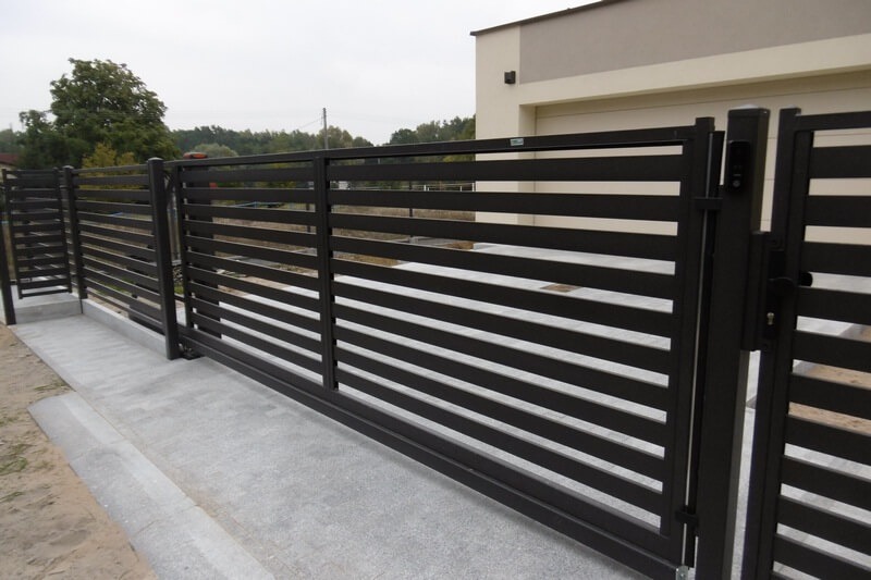 Nowoczesna, pozioma brama przesuwna w kolorze antracytowym, z furtką i domofonem, na betonowej wylewce przed domem z jasną elewacją.