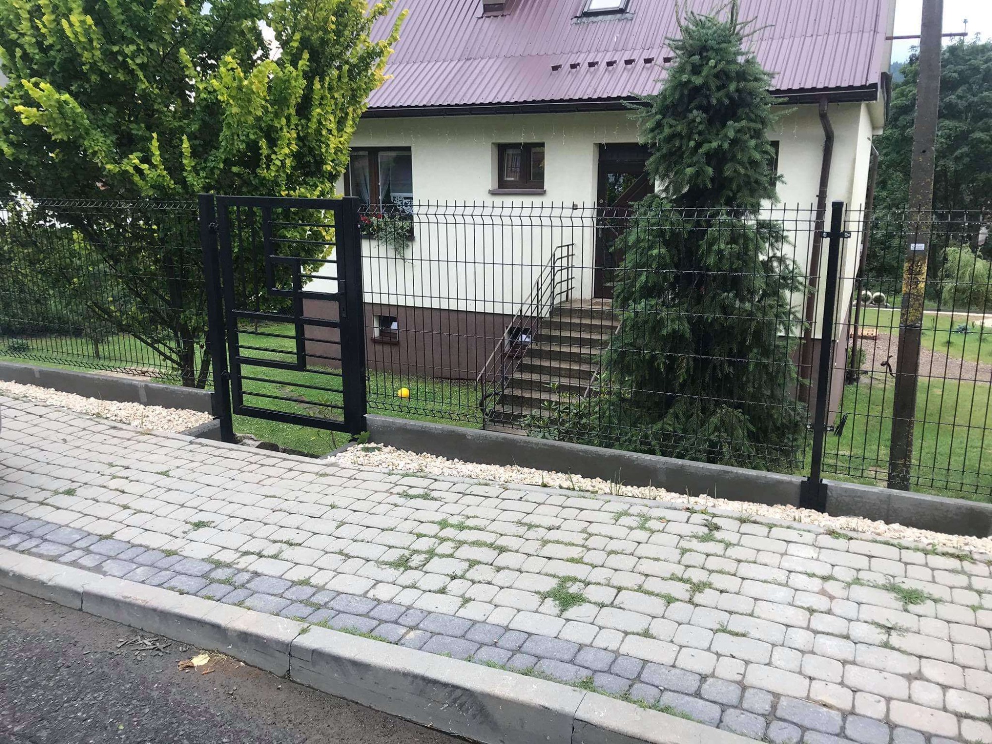 Nowoczesne, czarne ogrodzenie z paneli z furtką, oddzielające posesję od chodnika wyłożonego kostką brukową, z widokiem na dom z brązowym dachem i zielenią w tle.