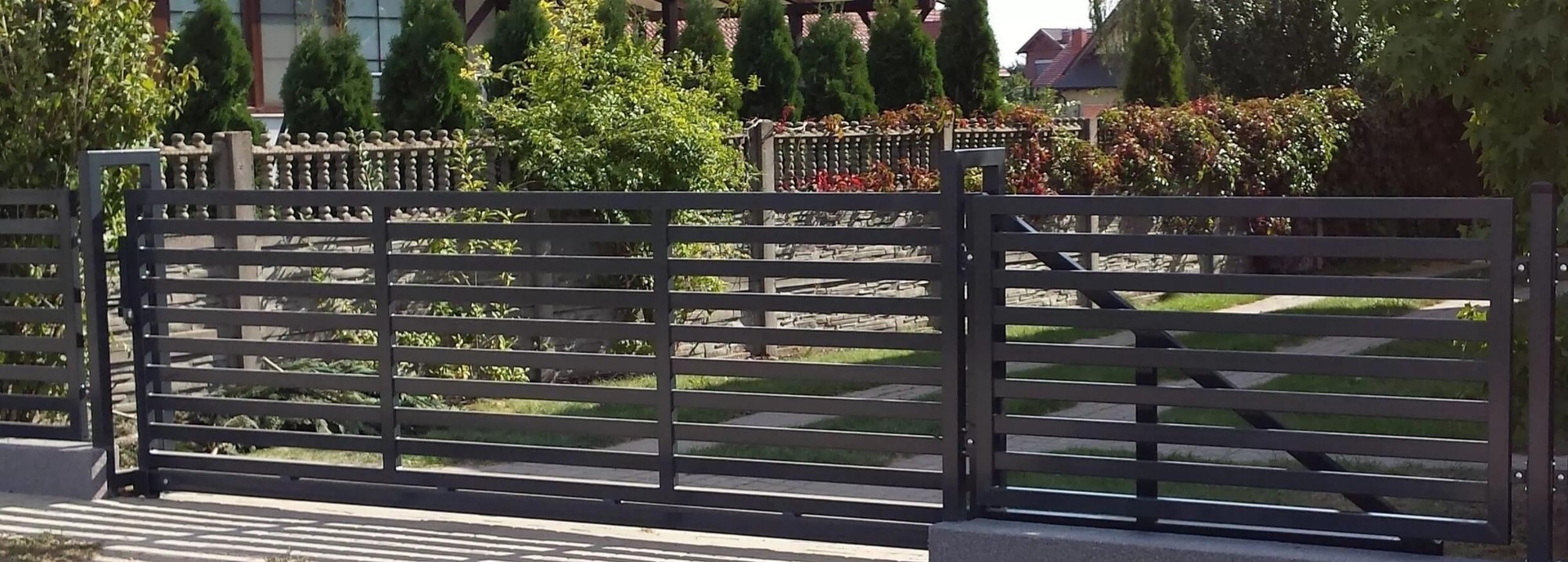 Nowoczesna, przesuwna brama metalowa z poziomymi profilami, w tle zadbany ogród z tujami i kamiennym ogrodzeniem