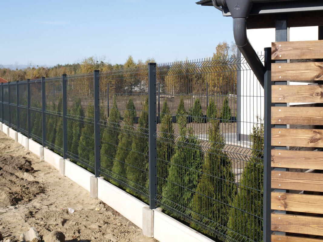 Nowoczesne ogrodzenie panelowe w kolorze grafitowym z podmurówką betonową i elementami drewnianymi, obsadzone tujami, widok na tle drzew.