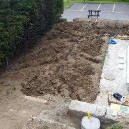 Przygotowany teren pod ogród z wykopanymi rowkami i częściowo ułożonymi kamiennymi schodami, widoczne niebieskie rury i zraszacze, w tle plac zabaw.