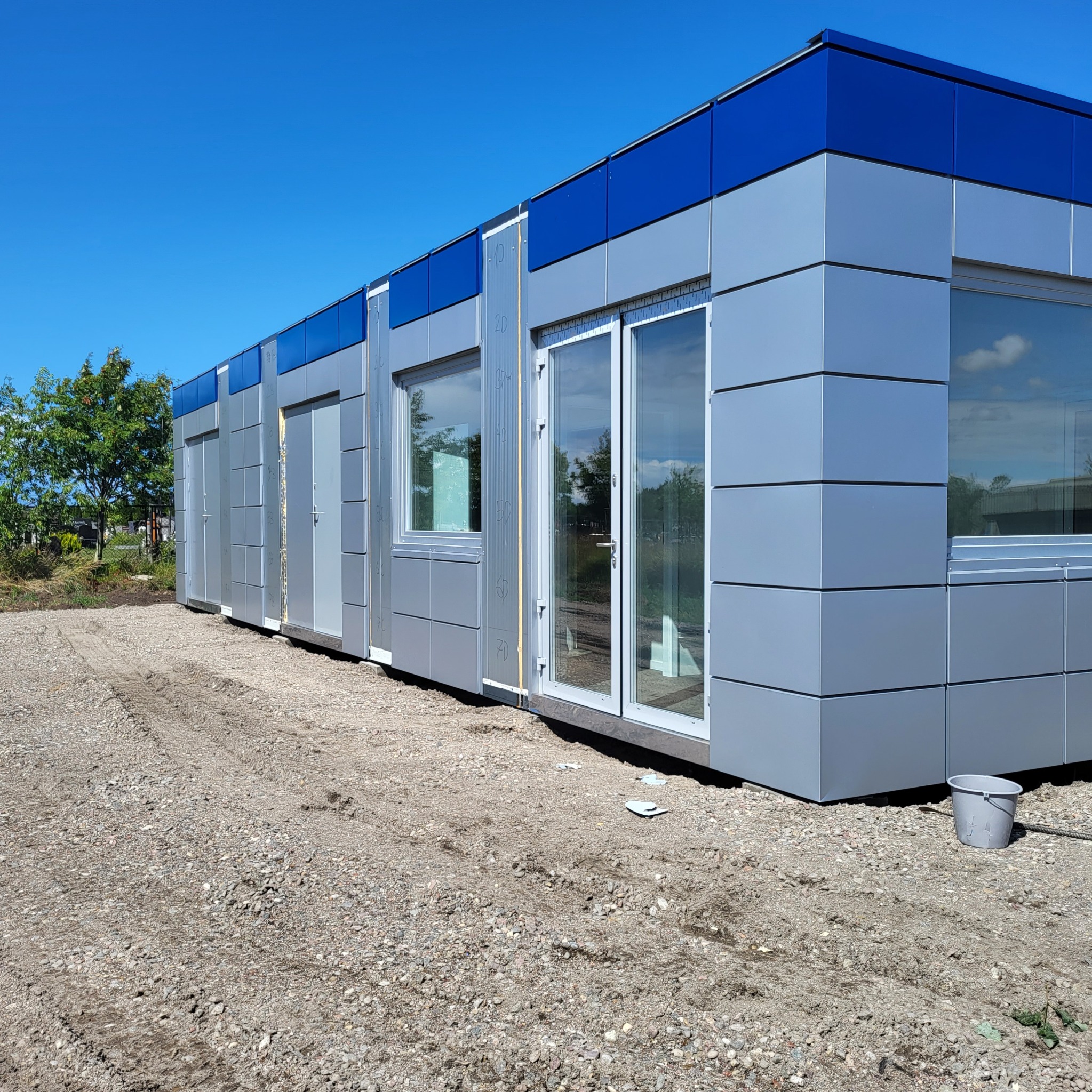 Modułowy budynek w kolorze szarym z niebieskimi akcentami, widoczne okna i drzwi, teren wokół wysypany żwirem, błękitne niebo w tle.