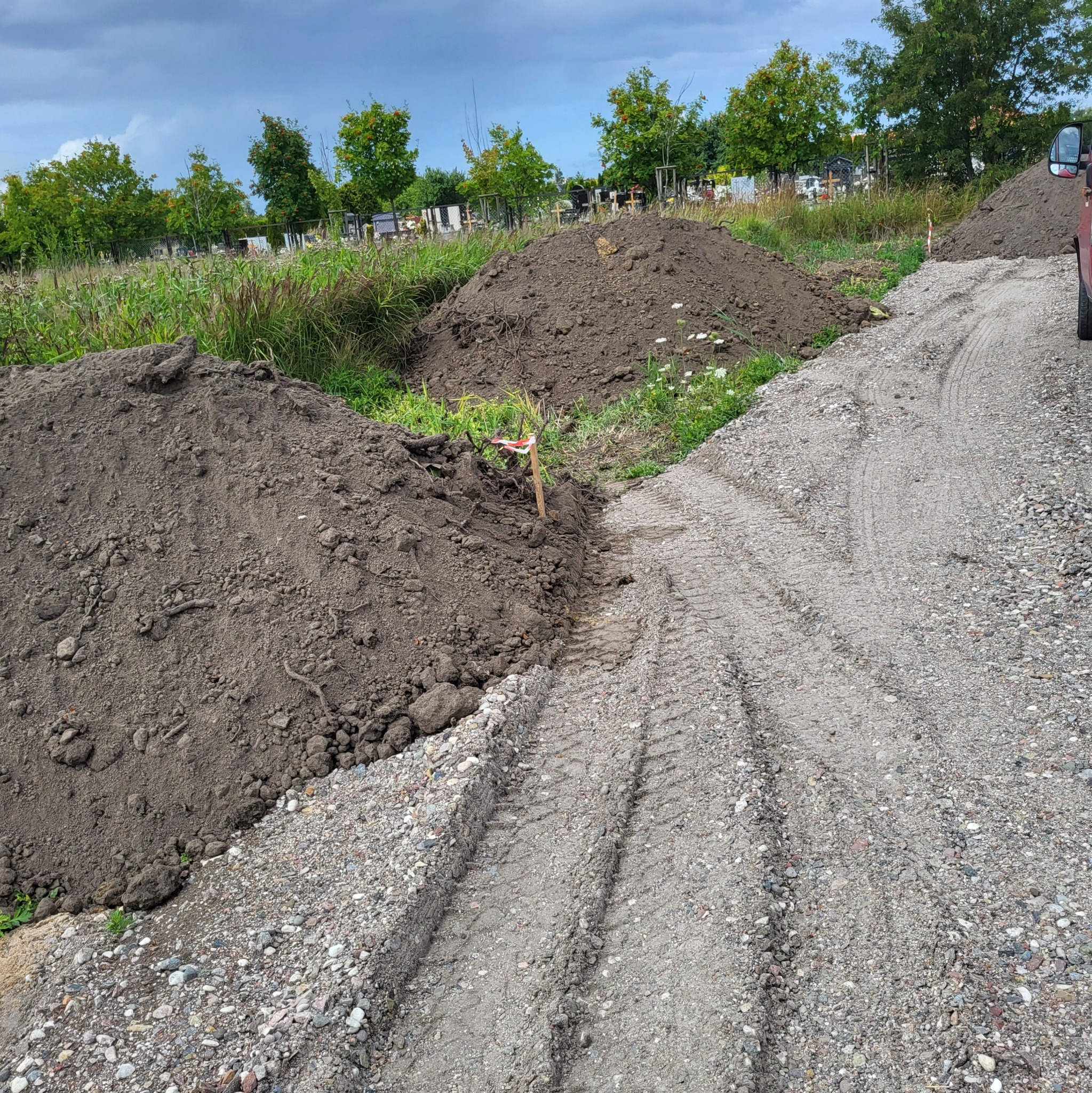Przygotowanie terenu pod przyszły ogród: pryzmy ziemi i utwardzona droga z widocznymi śladami opon, w tle zieleń i nagrobki cmentarza.