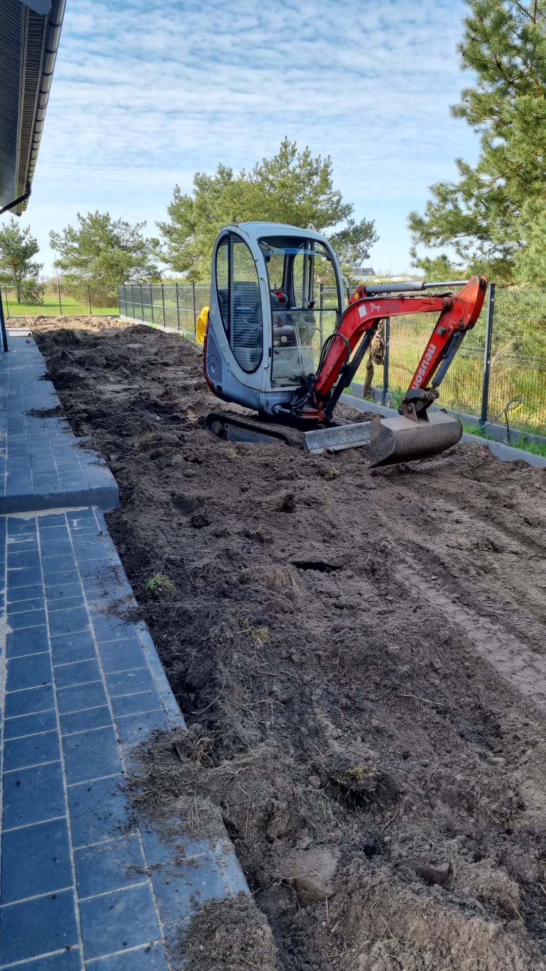 Czerwono-szara mini koparka Neuson na placu budowy, przygotowująca teren pod przyszły trawnik, widoczny fragment ułożonej kostki brukowej.